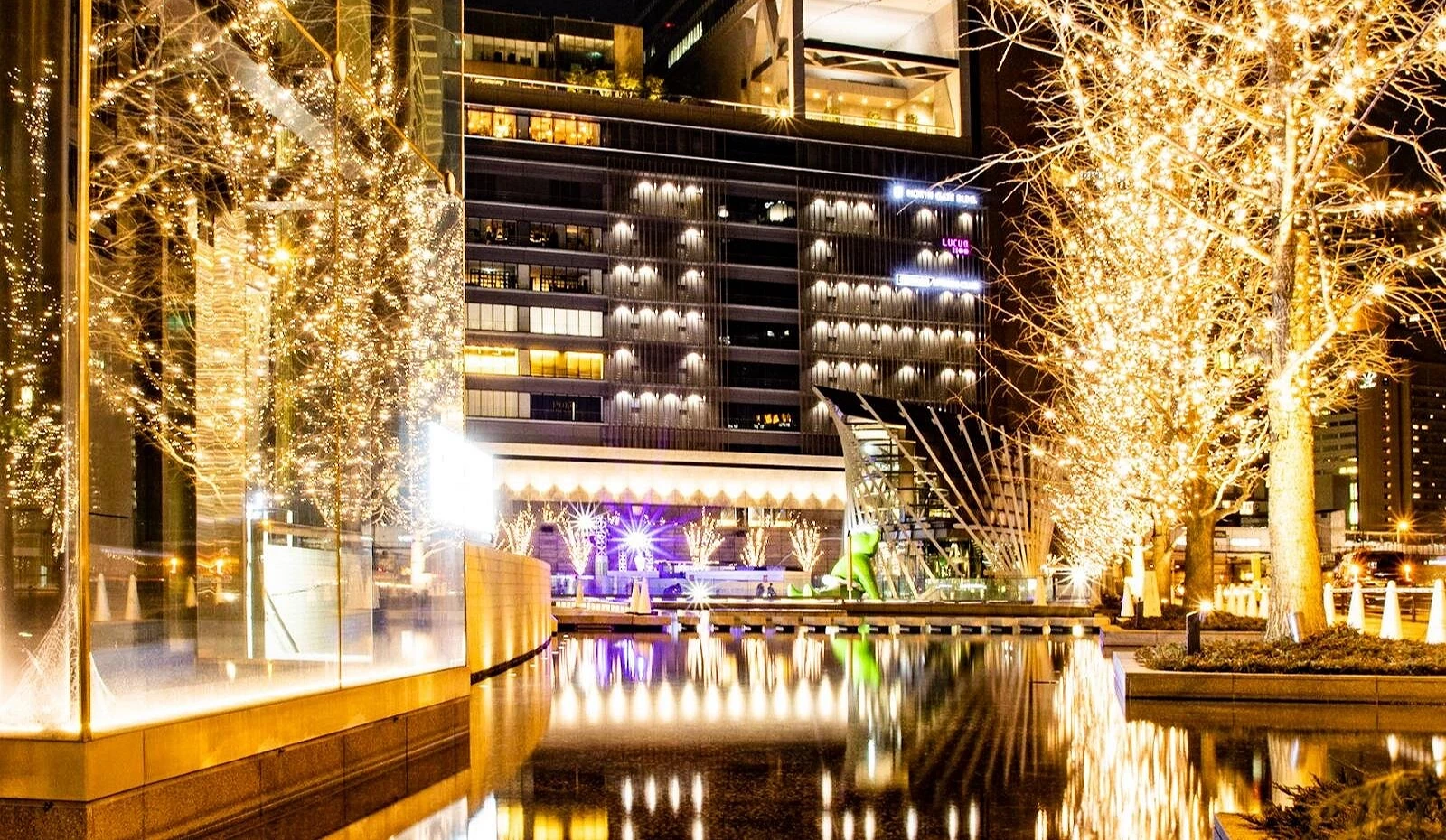 夜晚的 Grand Front Osaka 梅北广场被香槟金灯光包围,街树与建筑倒映在水池中,光影璀璨