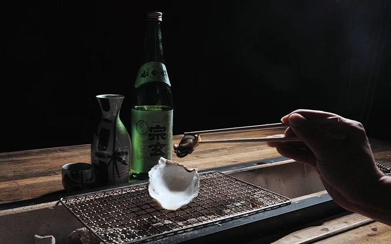 夾子將牡蠣從烤網上取起,後方放有「宗玄」清酒瓶