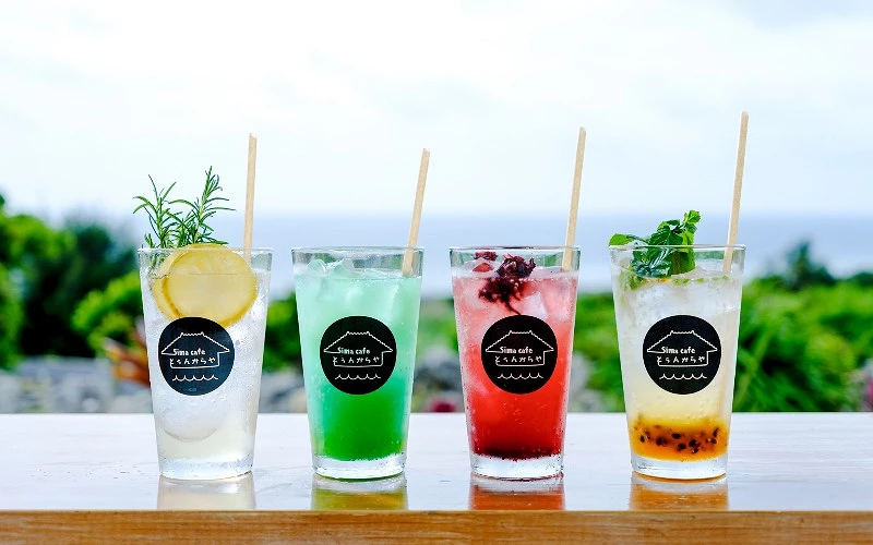 四杯彩色氣泡飲並排放置，杯身貼有咖啡店標誌，背景為海景