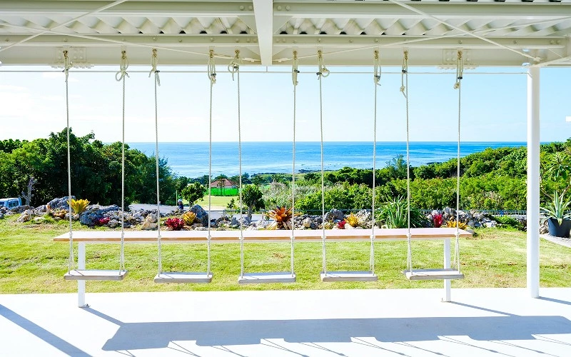戶外白色棚架下設置多座鞦韆，前方可眺望草地與海洋