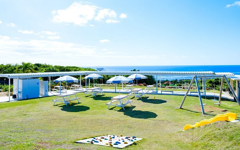 草地上設有野餐桌、遮陽傘與遊樂設施，背景可見海景