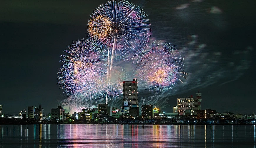城市煙火大會夜景,彩色煙火在高樓上空綻放,水面映照城市燈光與煙火倒影
