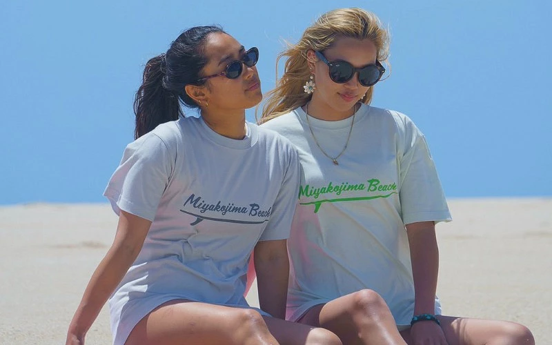 兩名女子坐在沙灘上，穿著印有 Miyakojima Beach 字樣上衣