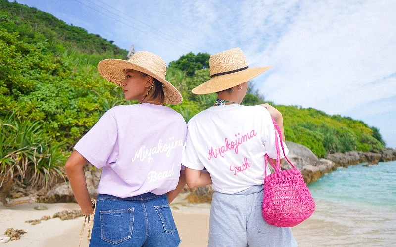 兩名女子背對鏡頭站在海邊，上衣背面印有 Miyakojima Beach 字樣