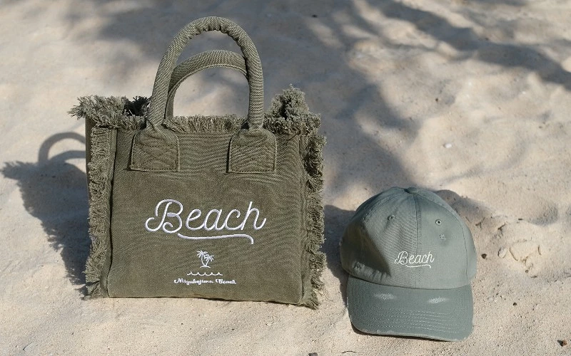 沙灘上擺放印有 Beach 字樣的帆布提袋與棒球帽