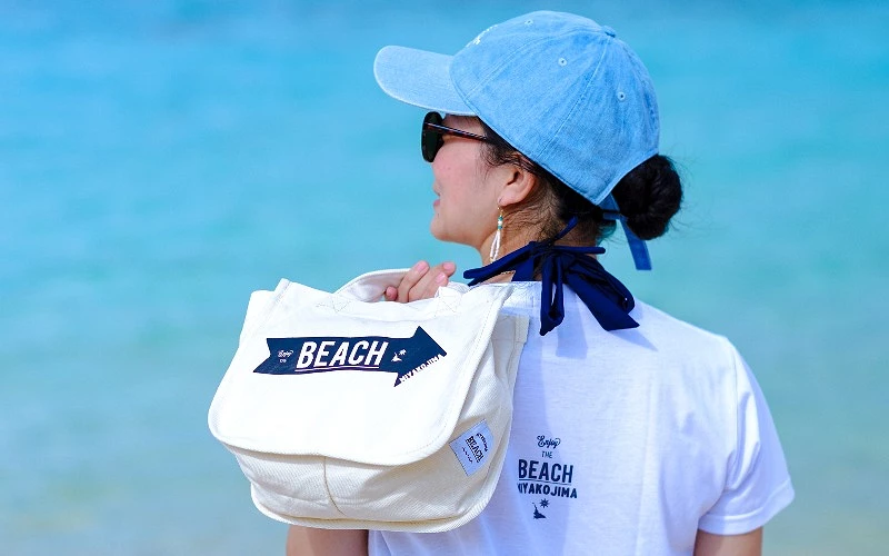 女子背對鏡頭站在海邊，肩背印有 BEACH 字樣白色帆布包