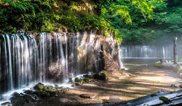 輕井澤白絲瀑布近景,陽光穿過樹林照在岩壁與水面上,細長水流沿岩壁流下,地面覆有石頭與淺水水潭