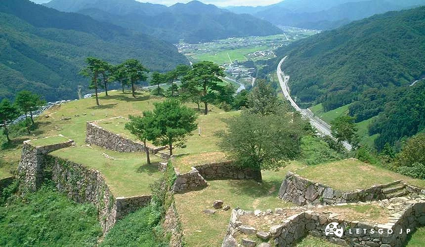 山丘上层叠石垣城迹遗址,散布松树,远眺山谷村落与蜿蜒道路,为竹田城迹