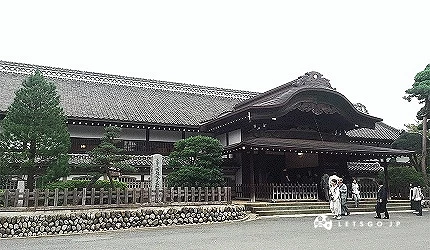 传统日式大型木造建筑前,数名身着和服人士伫立,周围有松树与石墙
