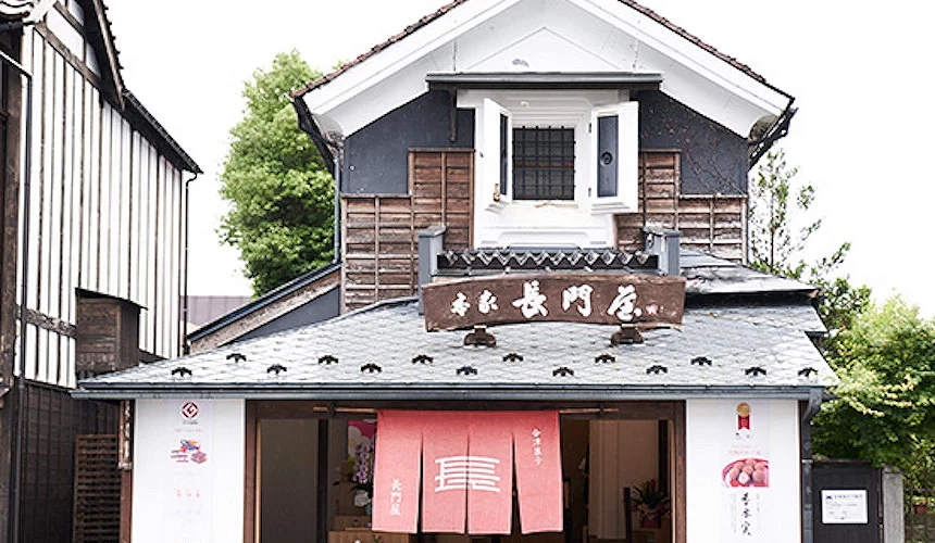 日式古民居风格店铺,白色下层搭配深蓝色上层,屋顶装饰瓦片,门口悬挂红色布帘和木制招牌写有日文。