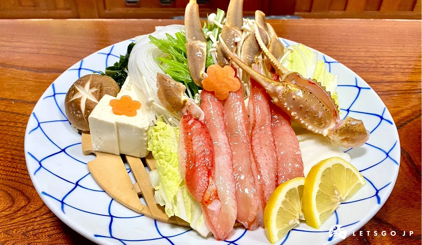一張特寫照片，白藍花紋的圓盤上整齊擺放著多支肉質紅潤、已去殼的長蟹腳，旁邊配有新鮮白菜、豆腐、香菇、金針菇與檸檬片。