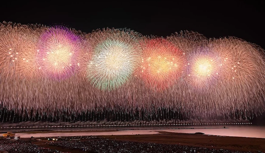 超寬幅煙火瀑布景觀,多組粉紅、金色、綠色大型煙火橫跨整個夜空,下方為海岸線及觀眾