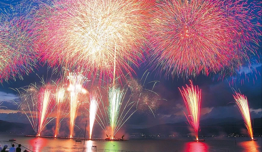 湖面上的煙火表演,金色、粉紅與白色煙火自水面發射升空,倒影映照於水面,岸邊有觀眾觀賞