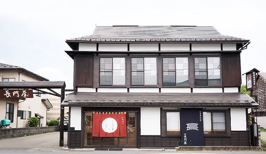 日式传统建筑店面,白黑色木造二层楼,门口悬挂红白圆形和深蓝色布幔,侧边招牌写有日文。
