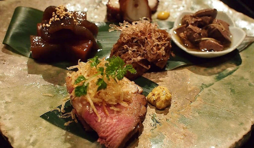 京料理餐厅「芜屋」的精致五种盛合拼盘,石盘上摆放着包含烤鸭肉、腌渍蔬菜等京都传统常备菜(Obanzai),背景为日式怀石餐饮氛围。