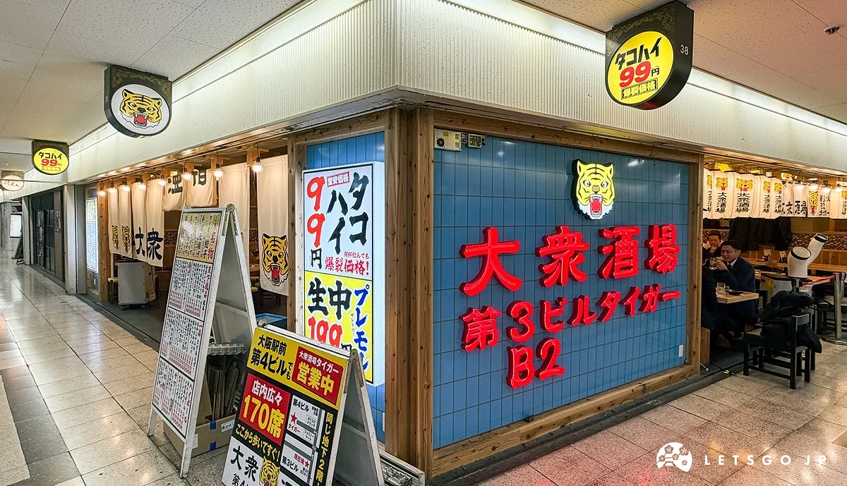 店鋪轉角處。藍色瓷磚牆面上貼著醒目的紅底黃字海報,寫著「大眾酒場 B2」。右側店內有穿著深色衣物的客人在桌邊用餐。
