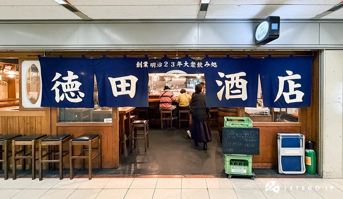 店面外觀。黑色木框架門面,上方掛著藍色橫向暖簾,印有白色漢字「徳田酒店」。門口右下方擺放著幾個塑膠啤酒筐