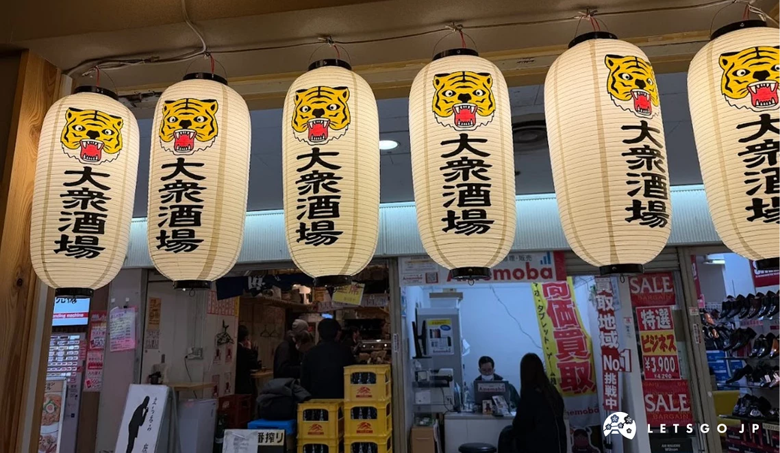 店頭懸掛的一排六個白色圓柱燈籠。燈籠上印有黑色老虎頭圖樣與「大眾酒場」四個漢字,內部透出黃色光線。