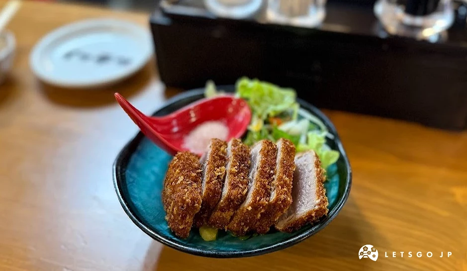 一個藍色瓷碗盛裝切塊炸鮪魚排。魚排外皮為深金黃色麵包粉,中心截面呈粉紅色半熟狀。後方附有生菜葉與一球白色大根泥。