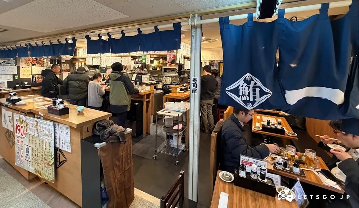 居酒屋內部,多位穿著深色西裝的男子站立於木製吧台前用餐。吧台上方掛著印有白色圖案的深藍色短簾,牆面上貼滿白色長方形的手寫菜單。