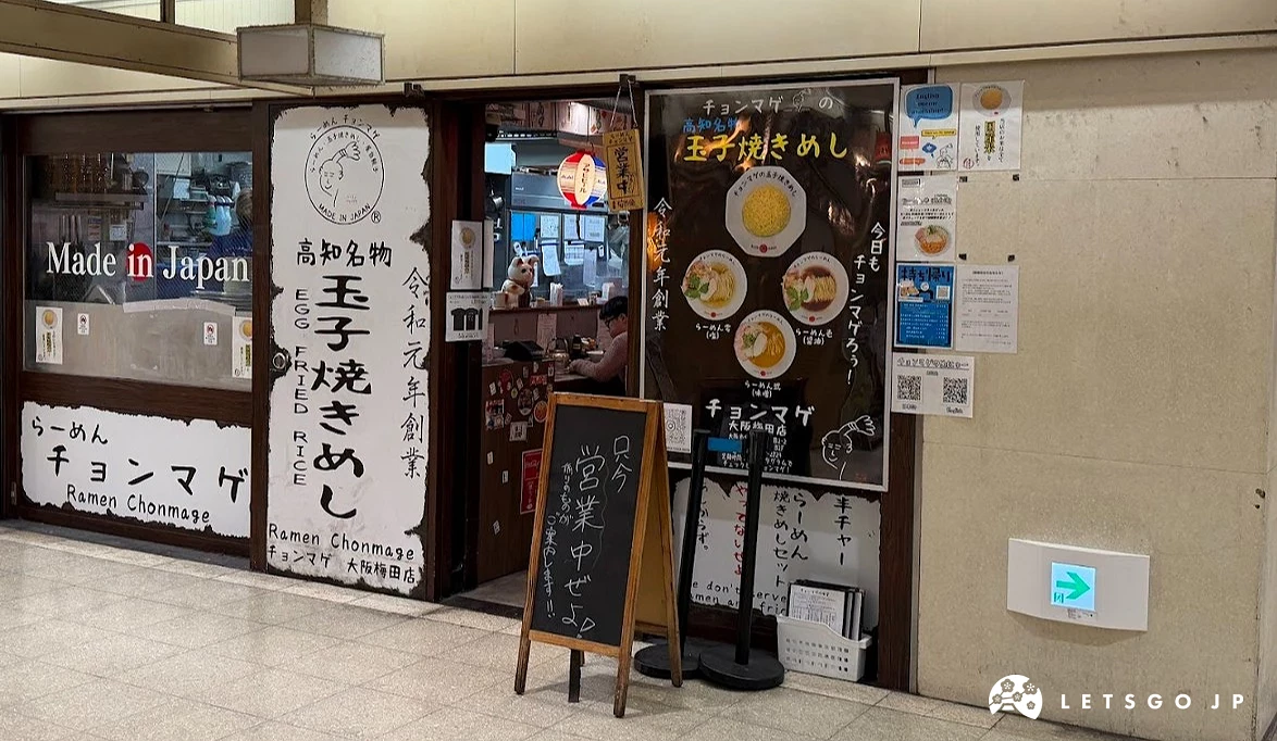 店門口掛有白色燈箱招牌,門旁張貼大型食物海報,海報中有一碗拉麵與一盤圓頂狀炒飯。門前立著一個黑色的小型 A 字型手寫菜單看板。