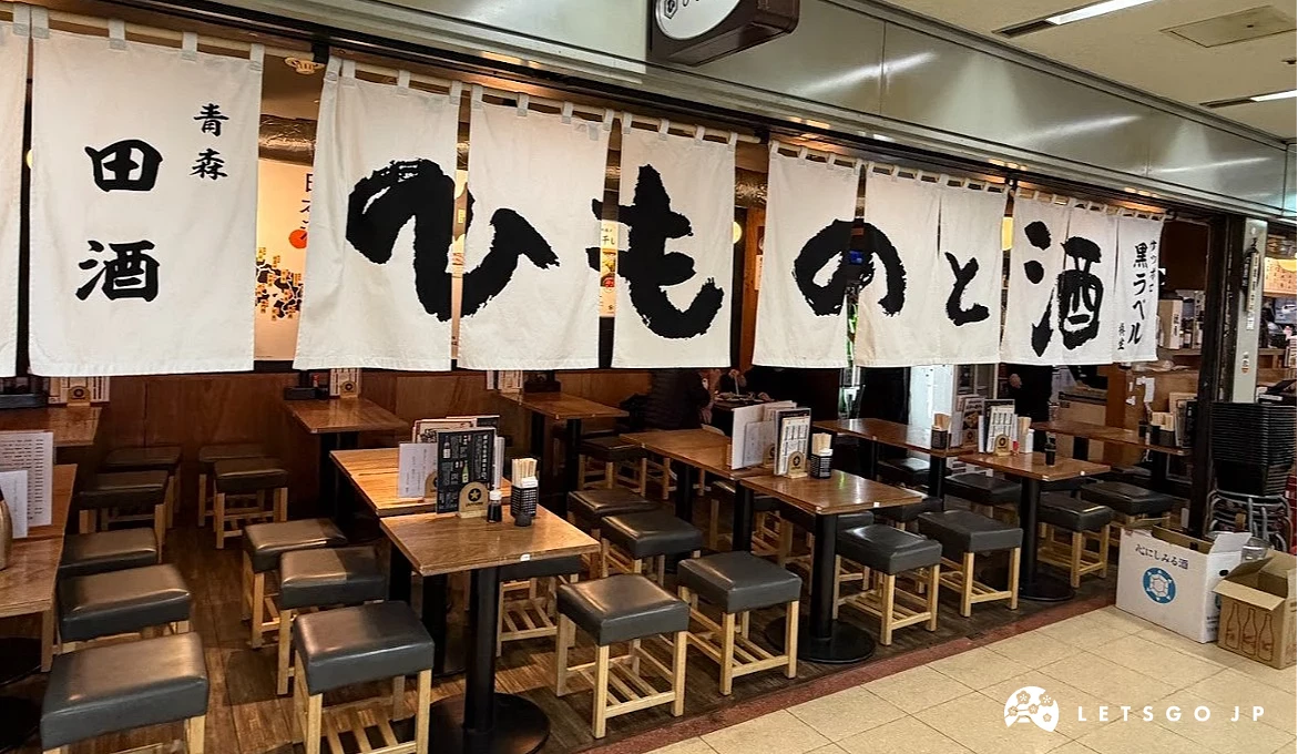 店內用餐區。多張木質長方形餐桌搭配黑色方形高腳凳。天花板掛著大面白色布簾,上面印有黑色的「ひものと酒」(魚乾與酒)字樣。
