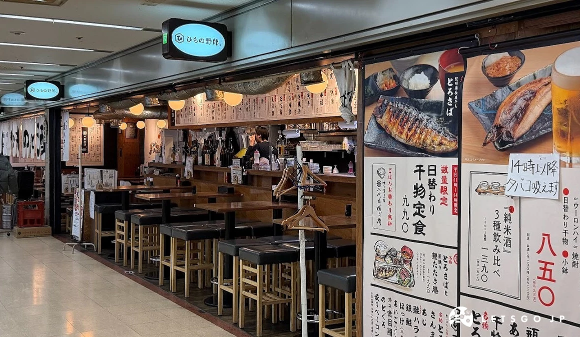 店面外牆貼滿了白色海報,海報上有烤魚照片與餐點價格。右側海報寫著「平日限定 850 日圓」。櫃檯內部有兩名工作人員,天花板掛著圓形的藍色小招牌。