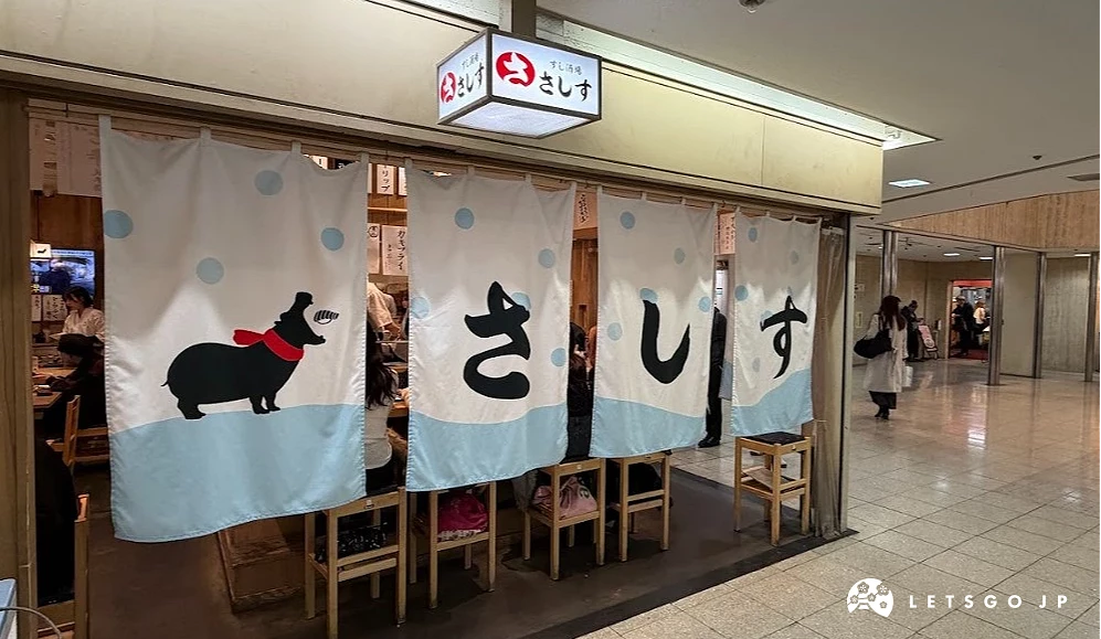 壽司店門面。上方掛著白色長暖簾,印有黑色臘腸狗插畫與藍色字體「さしす」。店內可見木質餐桌與多張木椅子。