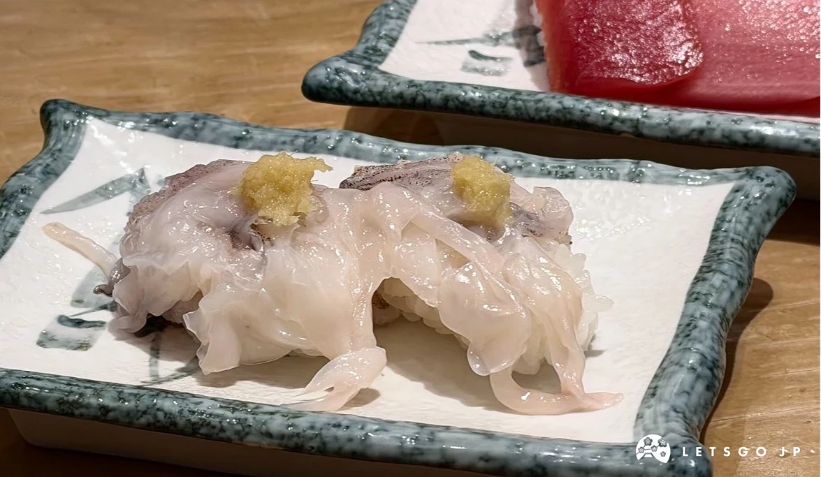 近距拍攝一盤白身魚壽司。魚肉呈半透明乳白色,上方放置少量薑泥與細蔥。後方背景有一盤深紅色的鮪魚生魚片。