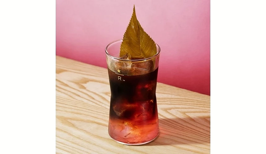 一杯樱花桶陈冷萃咖啡放在木桌上,饮品呈现深咖啡与粉红渐层,上方以樱叶装饰,玻璃杯印有星巴克臻选标志