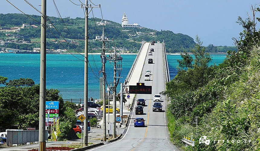 白色拱橋跨越翠藍海灣,車流穿梭其上,遠方有山景