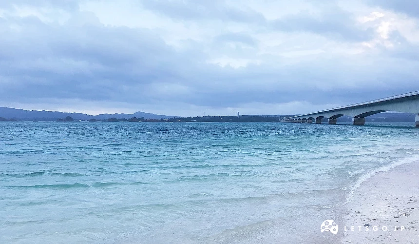 白沙灘浪花拍岸,長橋跨越碧藍海灣,遠山與雲影映襯寧靜海濱。