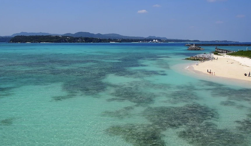 白沙灘遊客戲水,翠綠淺海清澈見底,遠山與船隻點綴海灣風景。