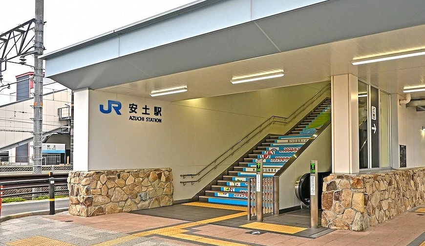 JR 安土駅站体入口正面,墙面写有「JR 安土駅 AZUCHI STATION」,楼梯踏面印有 2025 年活动彩绘图案,两侧为石砌墙面