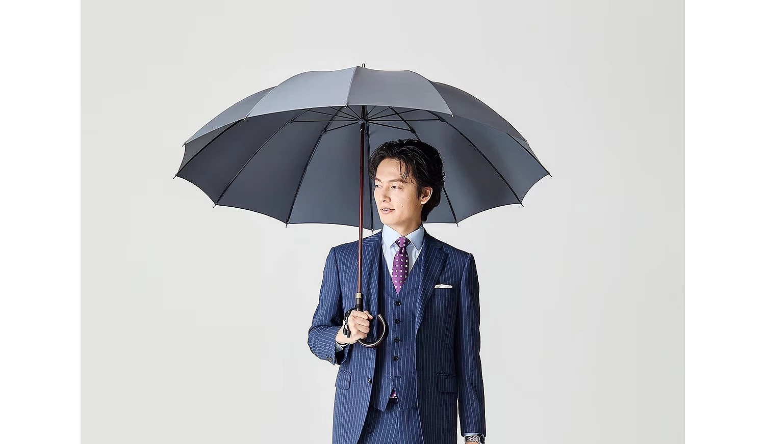 一名身穿深藍色條紋三件式西裝、搭配紫色圓點領帶的亞洲男子,手持一把深灰色木柄長傘,眼神看向側方