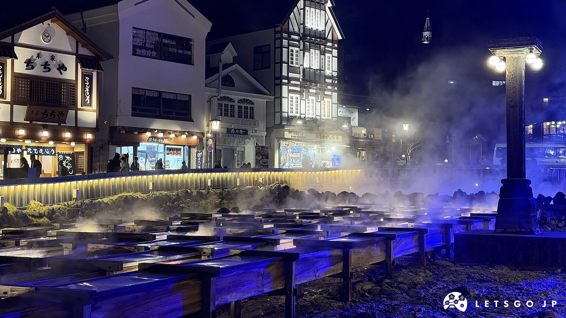 草津溫泉地標「湯畑」的夜景,前景為排列整齊的木製湯瀧,在藍色與白色燈光照射下,溫泉水蒸氣裊裊升起。