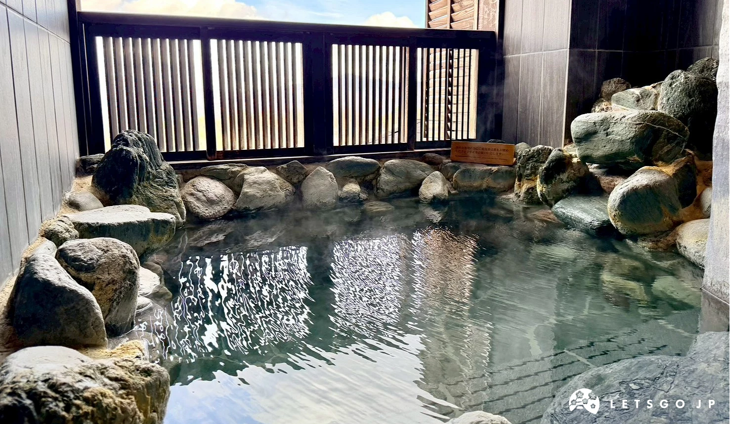 大型石造溫泉浴池，水面清澈，前方為廣闊的山丘景緻。