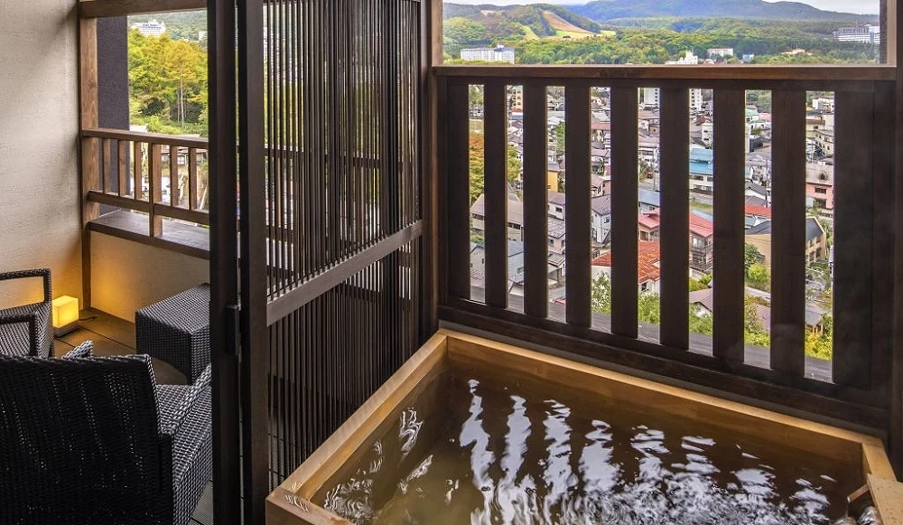 木質方型溫泉浴池，水流正注入池中，旁邊為設有戶外座椅的陽台空間