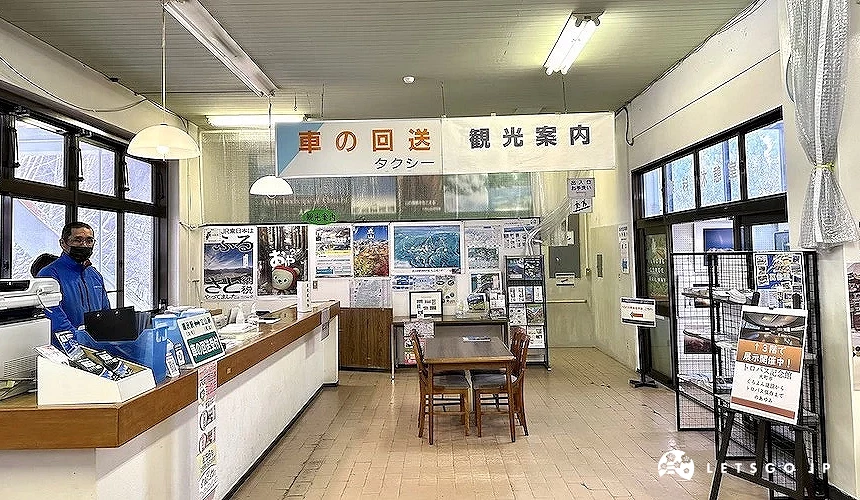 日式觀光案內所內部寬敞，職員立於木質櫃台，牆面貼滿寫有日文標語的旅遊資訊與風景照片。