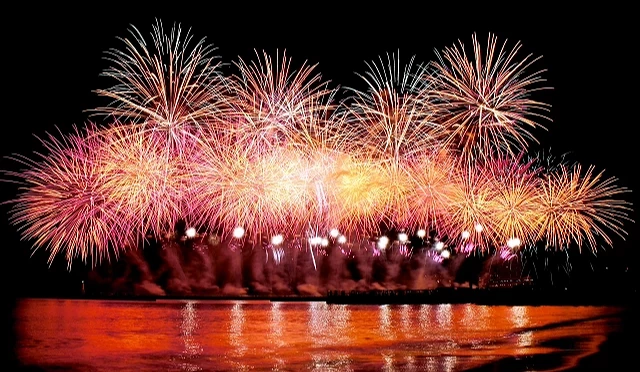 熱海海上花火大會於夜晚施放，大片金色與粉色煙火在海灣上空綻放，煙火光芒倒映在海面上