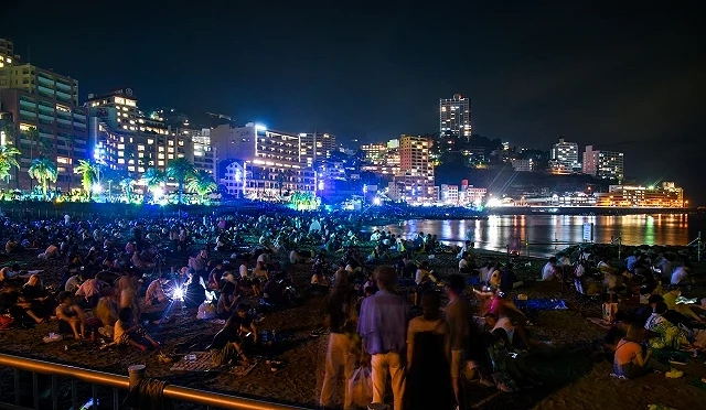 熱海花火大會夜晚會場人潮聚集，民眾坐在海灘上等待煙火施放，背景可見熱海市區與海灣夜景