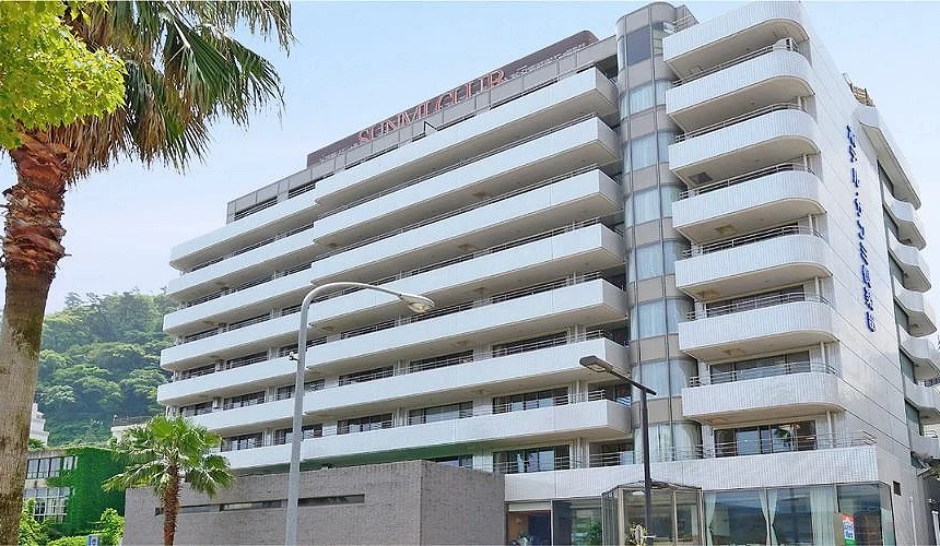 熱海海邊溫泉飯店外觀，白色建築設有多層陽台，前方有棕櫚樹，後方可見綠色山景