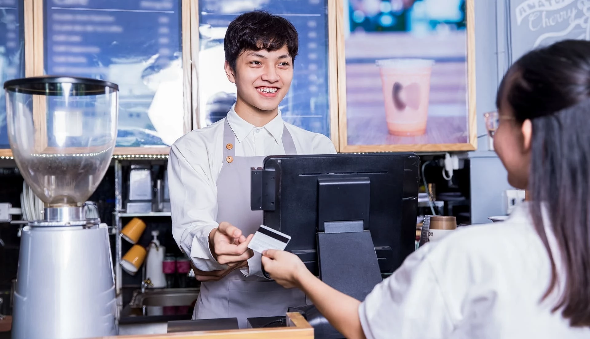 咖啡店柜台前,顾客递出信用卡付款,穿着围裙的店员在收银机后方微笑接待