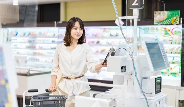 超市自助结帐区内,女性顾客推着购物篮,手持手机在付款机前进行电子支付