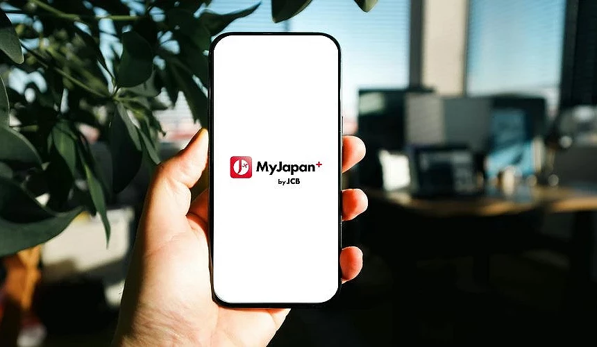 手持手机显示 MyJapan+ by JCB App 标志,背景为室内桌面与绿色植物,呈现日本旅游 App 使用情景