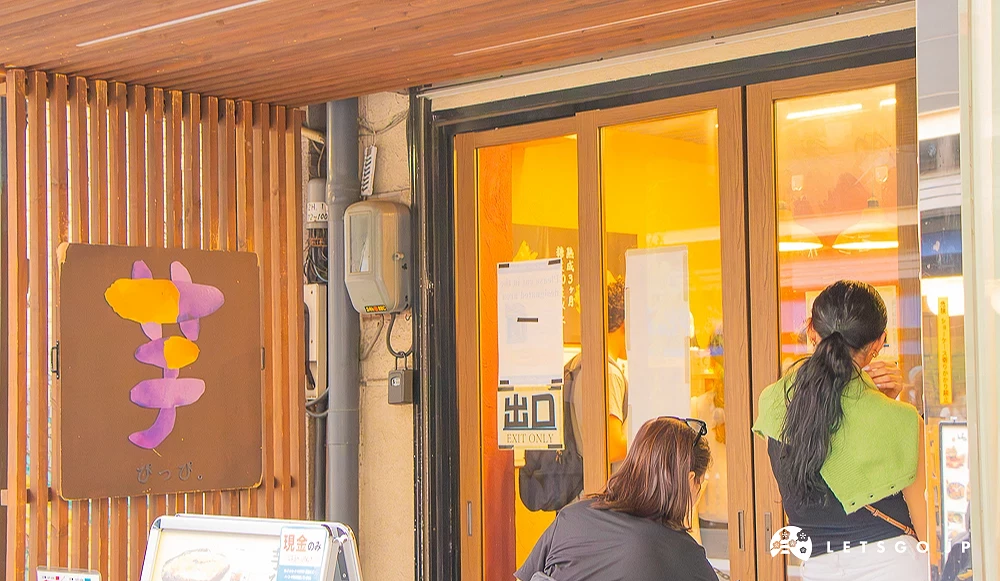 芋ぴっぴ。淺草店 「芋ぴっぴ。」地瓜甜點店門口,掛有紫色地瓜圖案招牌,遊客正於門外等待。