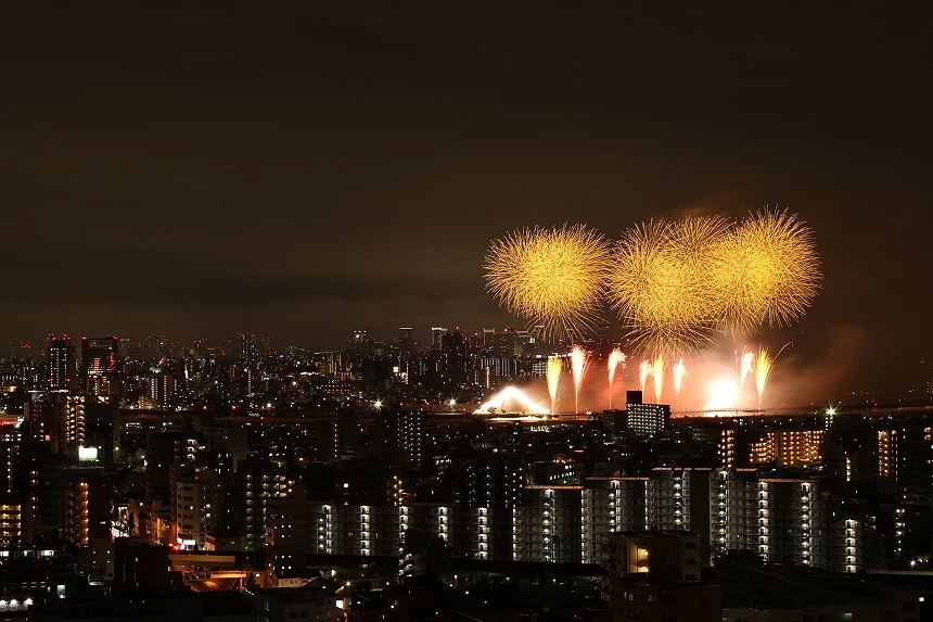 燈火通明的城市建築剪影上方,三組巨大的金黃色放射狀煙火在夜空中平行排列,映襯著下方的光束與煙霧。