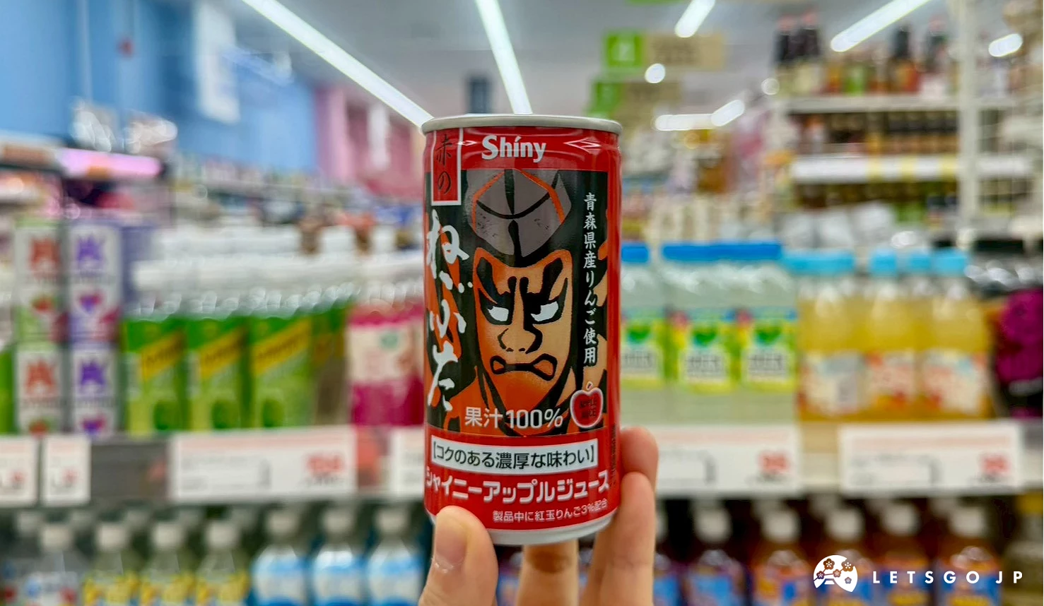一隻手拿著印有威武紅色睡魔祭面具圖騰的Shiny青森睡魔100%蘋果汁鐵罐裝,背景為藥妝店飲料架。