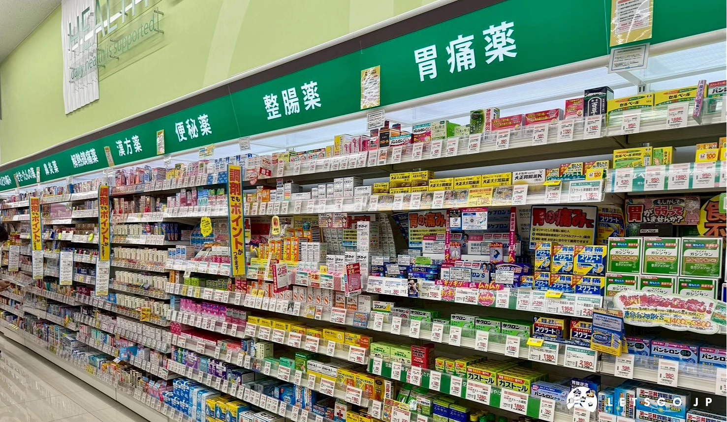 Happy Drug藥妝店內整齊排列的醫藥品陳列架,上方掛有綠色底、白色字的整腸藥與胃痛藥日文分類指標牌
