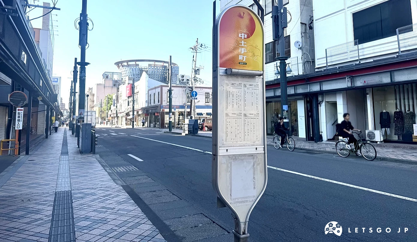 弘前市區街道旁的公車站牌,頂端圓形標誌印有黃底紅色蘋果圖案,下方標示著中土手町站名。